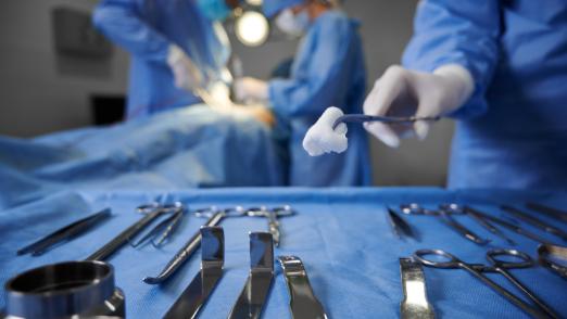 Close up of doctor hand holding forceps with tampon while surgeon and assistant performing plastic surgery on blurred background. Medical team doing cosmetic surgery in operating room at hospital.